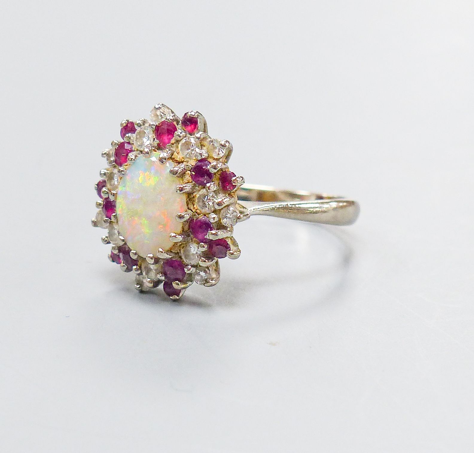 An 18ct white metal, white opal, ruby and diamond set oval cluster ring, size N, gross weight 6.1 grams.